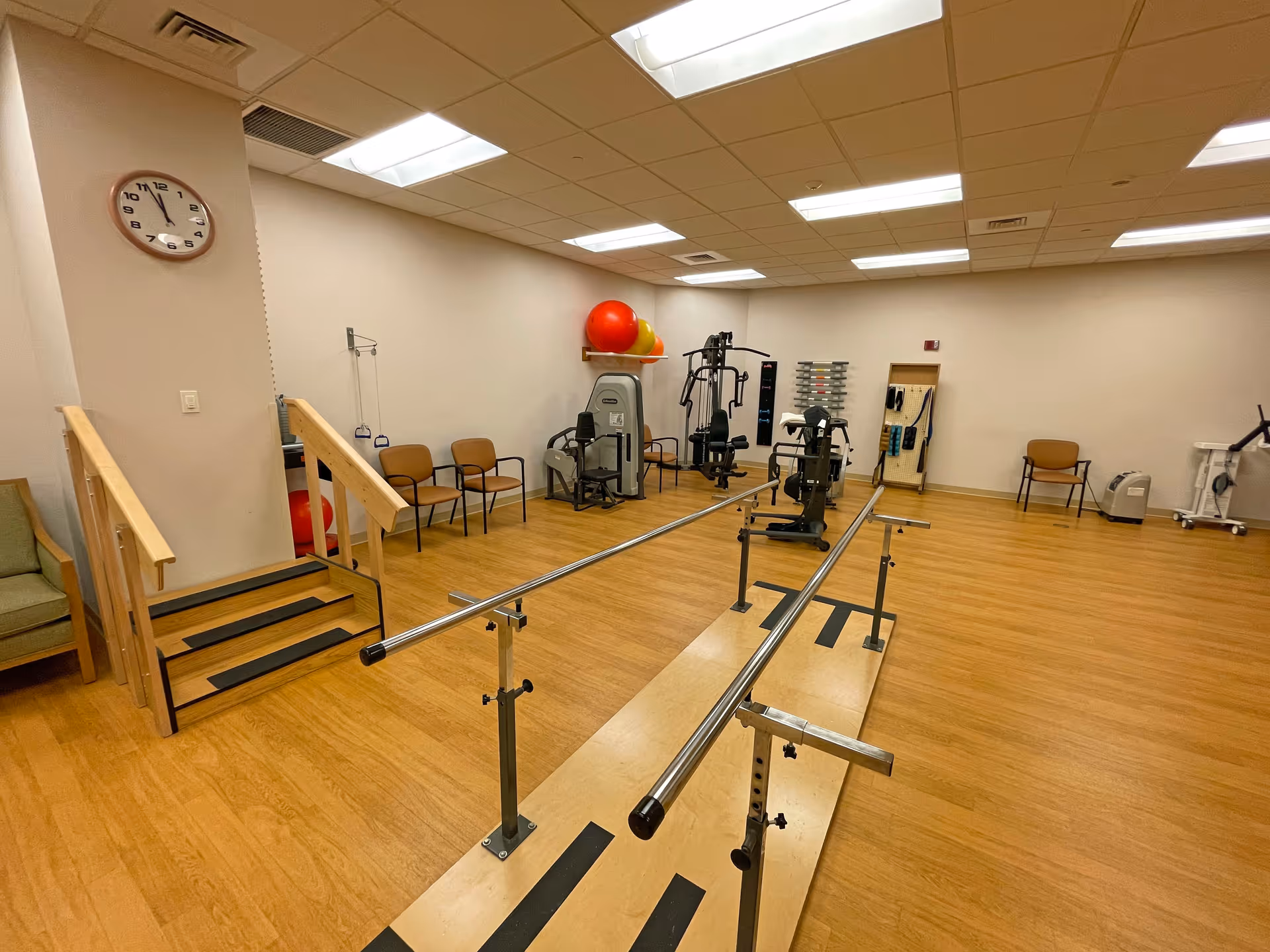 A physical therapy or rehabilitation room with wooden flooring, parallel bars for walking exercises, exercise machines, therapy balls on a shelf, chairs along the wall, and a small set of stairs with handrails. The room is well-lit with ceiling lights and has a clock on the wall.
