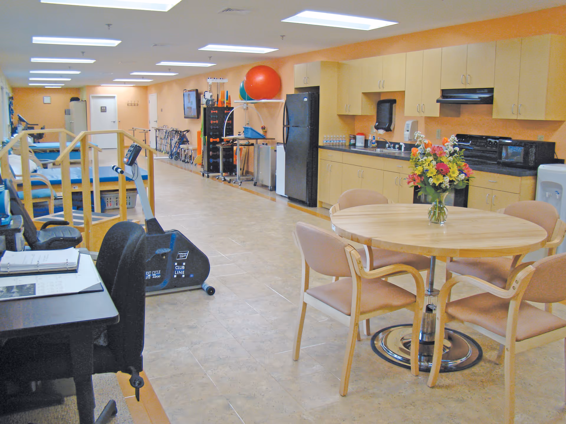 A spacious rehabilitation room with exercise equipment including stationary bikes and parallel bars. There is a round wooden table with four chairs and a vase of colorful flowers on it. The room has light-colored tiled flooring, beige walls, and a kitchenette area with cabinets, a refrigerator, microwave, and sink along one wall.