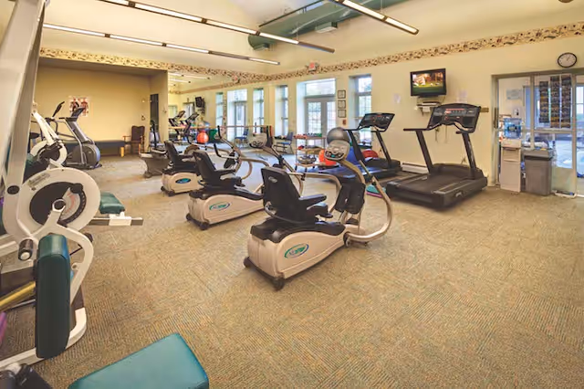 Bright fitness room with treadmills, recumbent exercise bikes, weight machines and large windows.