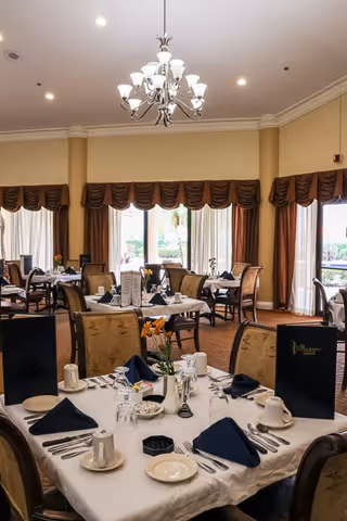 A well-lit dining room with multiple tables set for a meal. Each table is covered with a white tablecloth and set with plates, cups, silverware, and navy blue napkins. There are menus and small flower arrangements on the tables. Large windows with brown curtains allow natural light to fill the room, and a chandelier hangs from the ceiling.