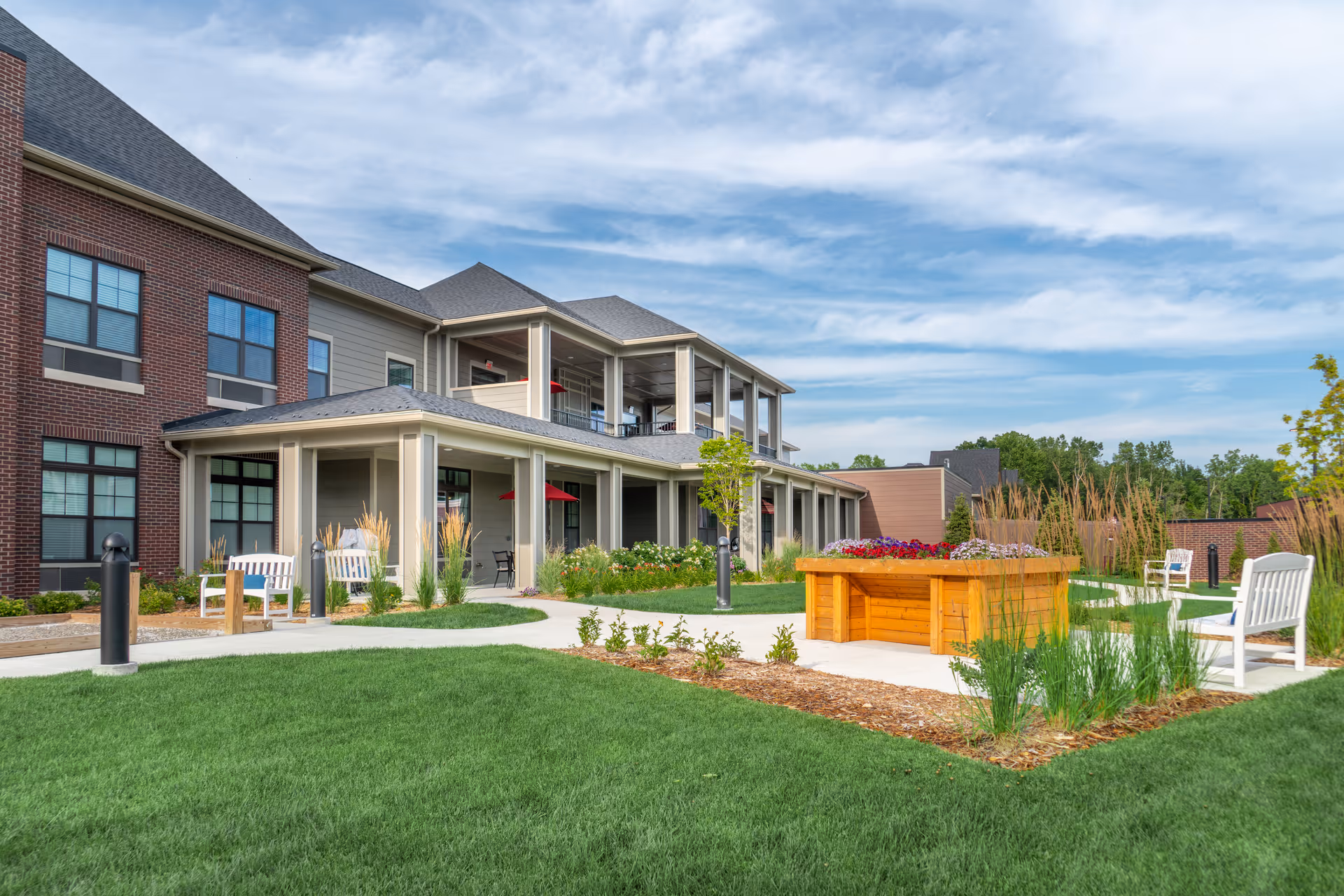 Outdoor view of a senior living facility with a well-maintained lawn, paved walkways, white benches, and a raised wooden flower bed filled with colorful flowers. The building has a combination of brick and siding exterior with large windows and covered porch areas.