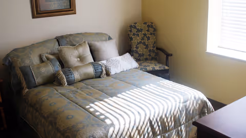 A bedroom with a neatly made bed featuring patterned bedding and multiple pillows. There is a cushioned armchair with a floral pattern next to the bed, and sunlight streams through a window with blinds, casting shadows on the bed. A framed picture hangs on the wall above the bed.