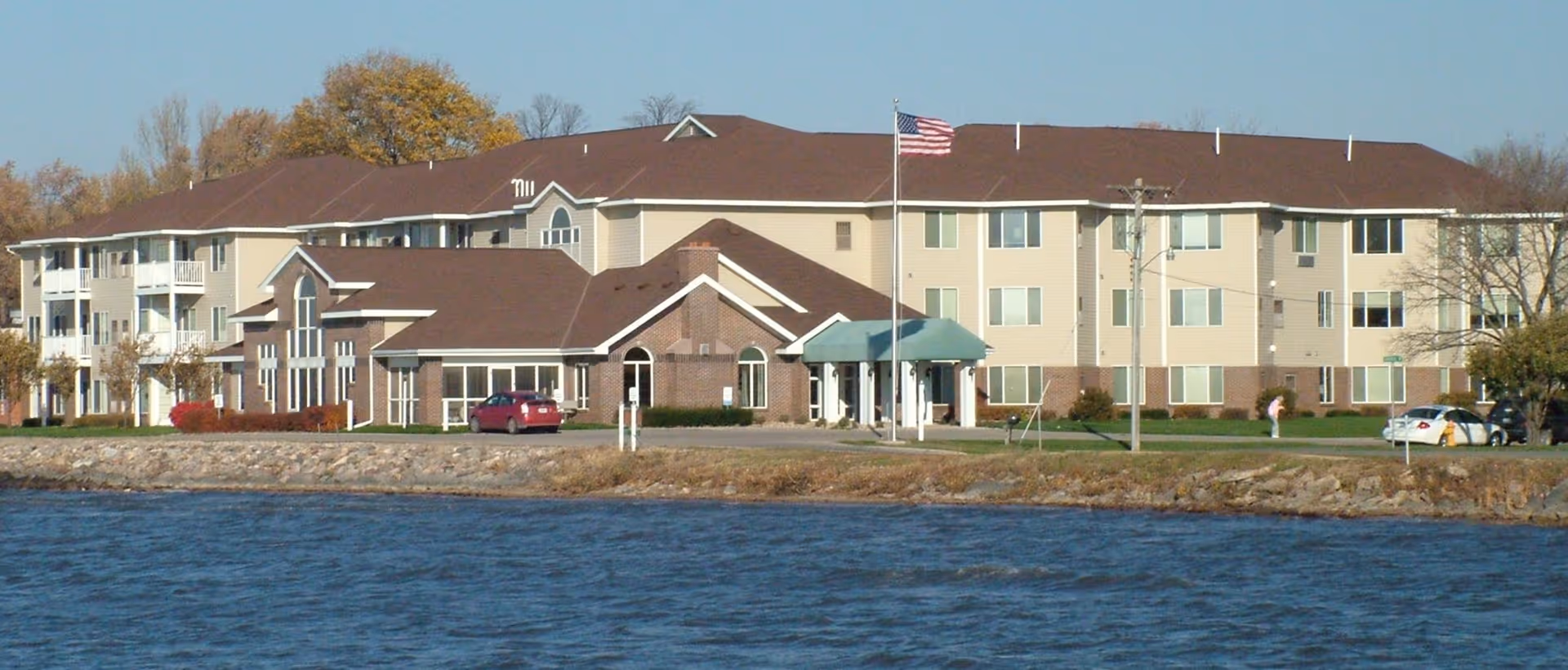 Exterior view of a large three-story senior living facility building with beige siding and brown roof, situated near a body of water. The building has multiple windows, a covered entrance with a flagpole flying the American flag, and some parked cars in front. Trees with autumn foliage are visible in the background.