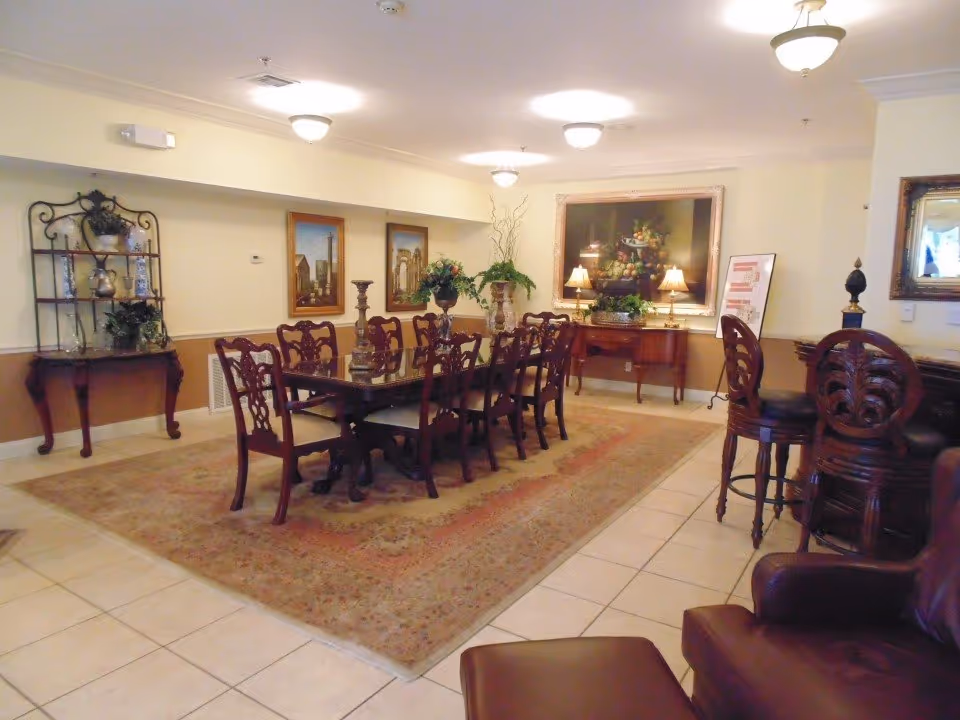 A formal dining room with a long wooden dining table surrounded by eight matching chairs. The room features a large decorative rug under the table, framed artwork on the walls, a sideboard with two lamps and a floral arrangement, and a small shelf with decorative items. There are also two high-backed bar stools near a counter on the right side and a leather armchair in the foreground.