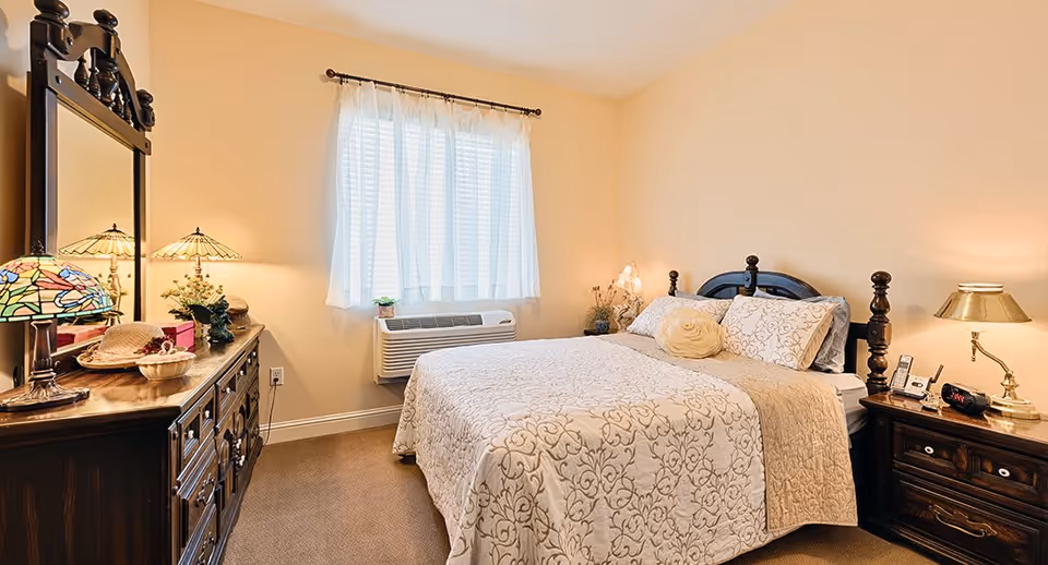 A cozy bedroom with a large bed featuring a patterned beige and white bedspread and multiple pillows. The room has a wooden dresser with a large mirror, decorative lamps, and various small items on top. A window with sheer white curtains allows natural light to brighten the room. There is a nightstand with a lamp, a clock, and a phone beside the bed. The walls are painted a soft beige color.