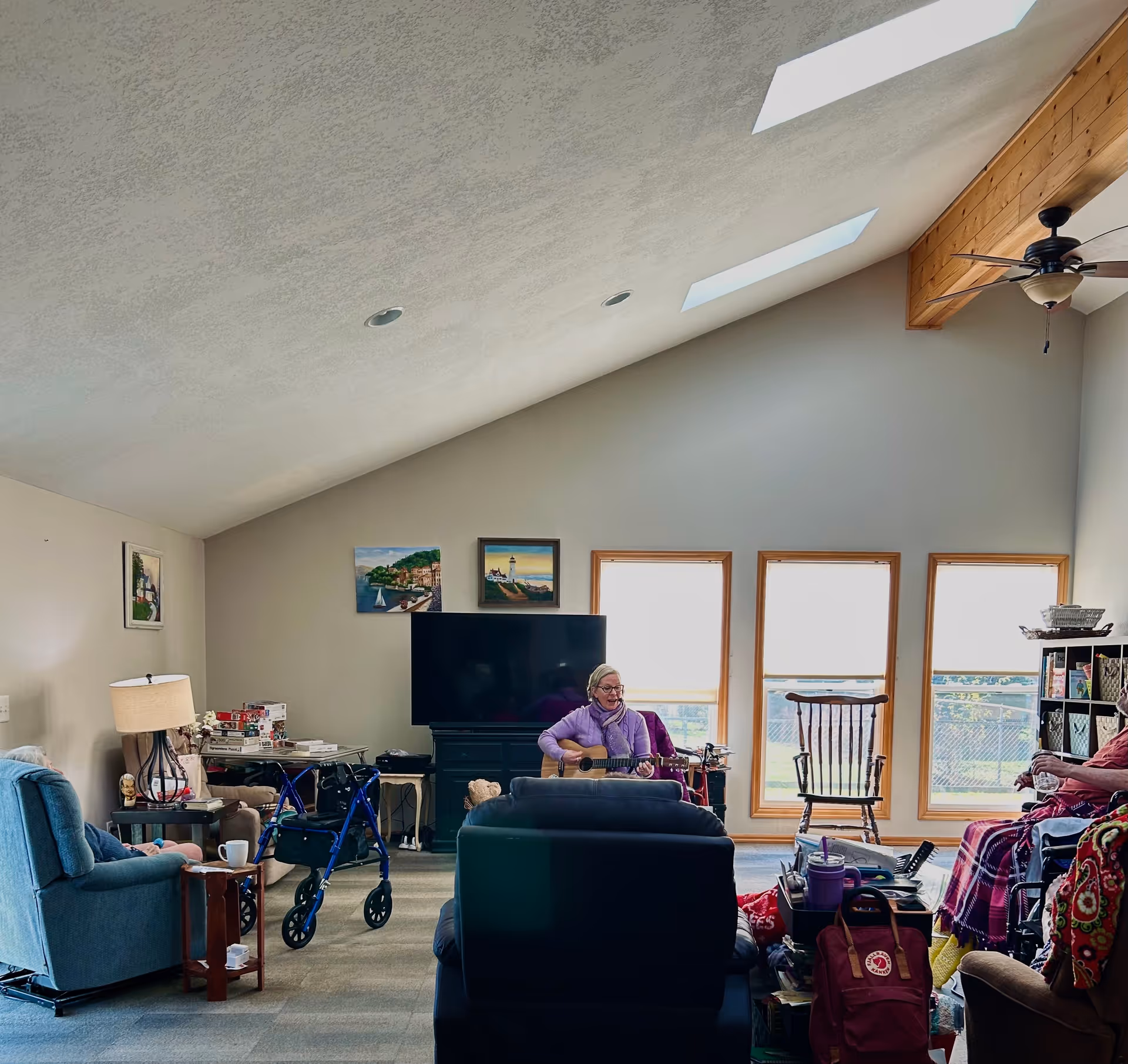 Vaulted living room with skylights, TV, seating and a resident playing a guitar near large windows.