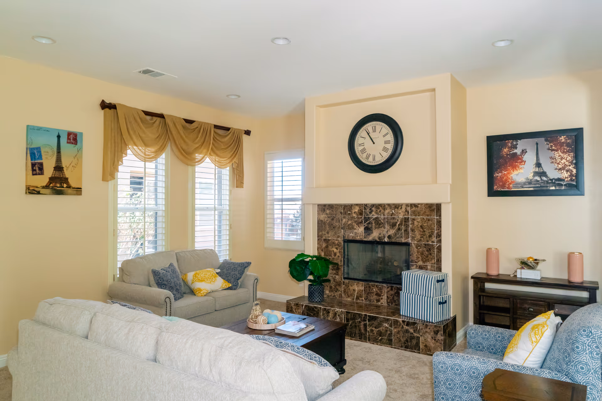 A cozy living room with beige walls and carpet, featuring a fireplace with a marble surround and a large clock above it. The room has two beige sofas and a patterned armchair with decorative pillows. There are three windows with white blinds and a yellow valance. Wall art includes two pictures of the Eiffel Tower. A wooden coffee table and a side table with decorative items complete the room.