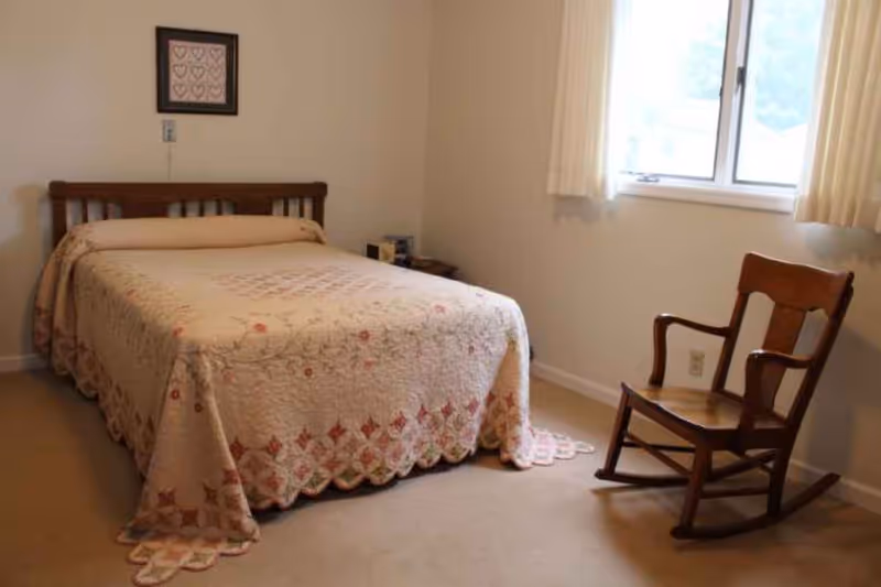 A simple bedroom with a wooden bed frame and a quilted bedspread featuring a floral pattern. There is a wooden rocking chair near a window with cream-colored curtains, and a small bedside table with some items on it. A framed picture with heart designs hangs on the wall above the bed.