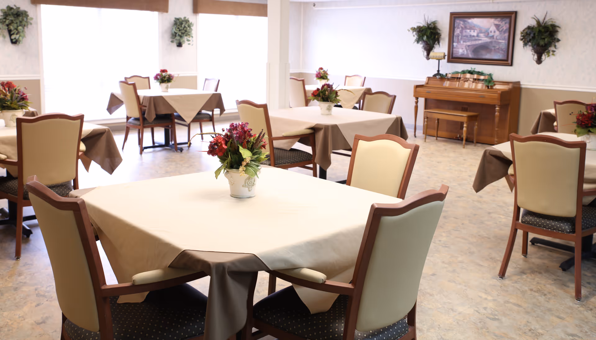 Dining room with multiple tables and chairs, each table covered with a cloth and a floral centerpiece, and a piano against the far wall.
