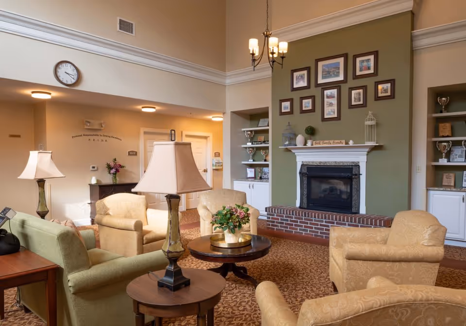 Cozy senior living common room with armchairs, side tables, lamps, and a fireplace topped with framed photos.