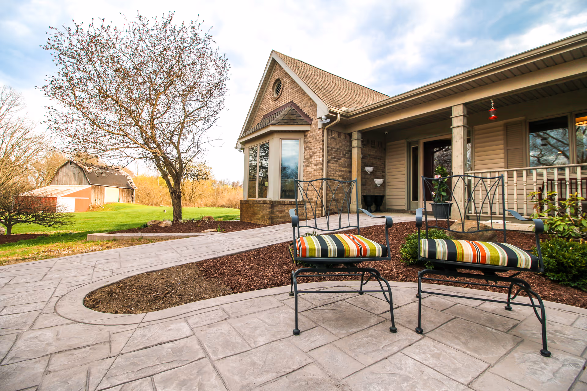 Outdoor patio area at TenderCare assisted living Oakland MI with two metal chairs featuring striped cushions, a paved walkway, a tree with sparse leaves, and a building with a porch and large windows in the background.