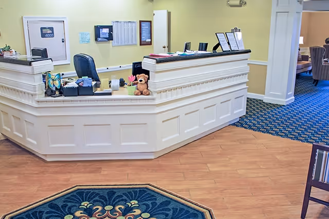 Reception desk area in a senior living facility lobby with a teddy bear on the counter and seating visible to the side.