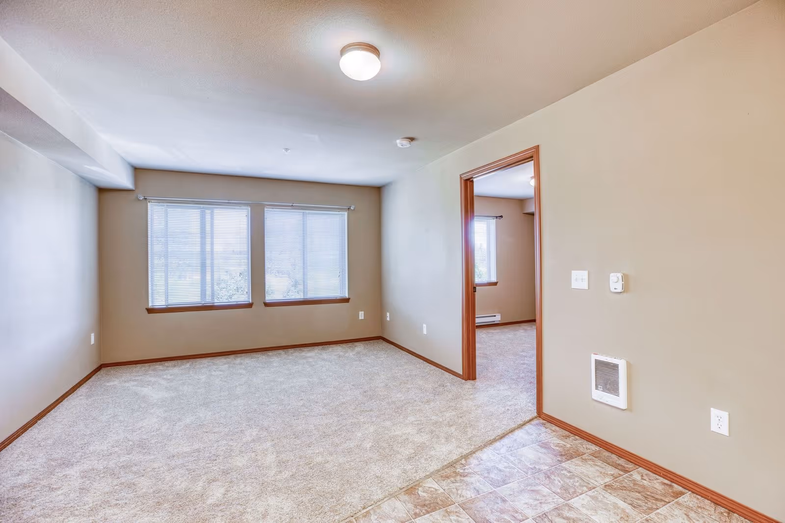 Empty room with beige walls and carpeted floor, featuring three large windows with blinds. Adjacent room visible through an open doorway, also carpeted with a window. The floor near the entrance is tiled, and there are electrical outlets and a wall heater on the right wall.