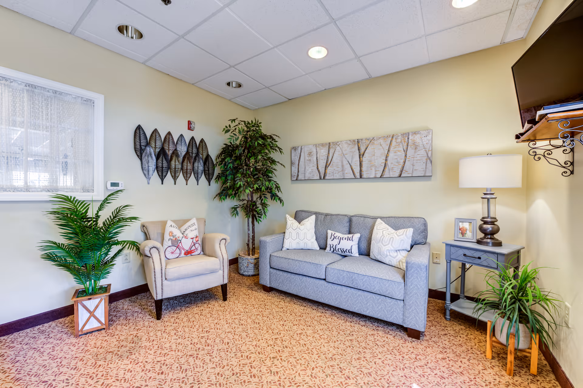 A cozy living room area with a light gray sofa adorned with three decorative pillows, including one that says 'Beyond Blessed.' Next to the sofa is a small gray side table with a lamp, a framed photo, and a green plant underneath. To the left of the sofa is a beige armchair with a decorative pillow featuring a bicycle design. The room has beige walls with a large artwork of birch trees above the sofa and metal leaf wall decor above the armchair. There are two potted plants on the floor, one near the armchair and one near the side table. A window with lace curtains is visible on the left wall, and a flat-screen TV is mounted on the right wall.