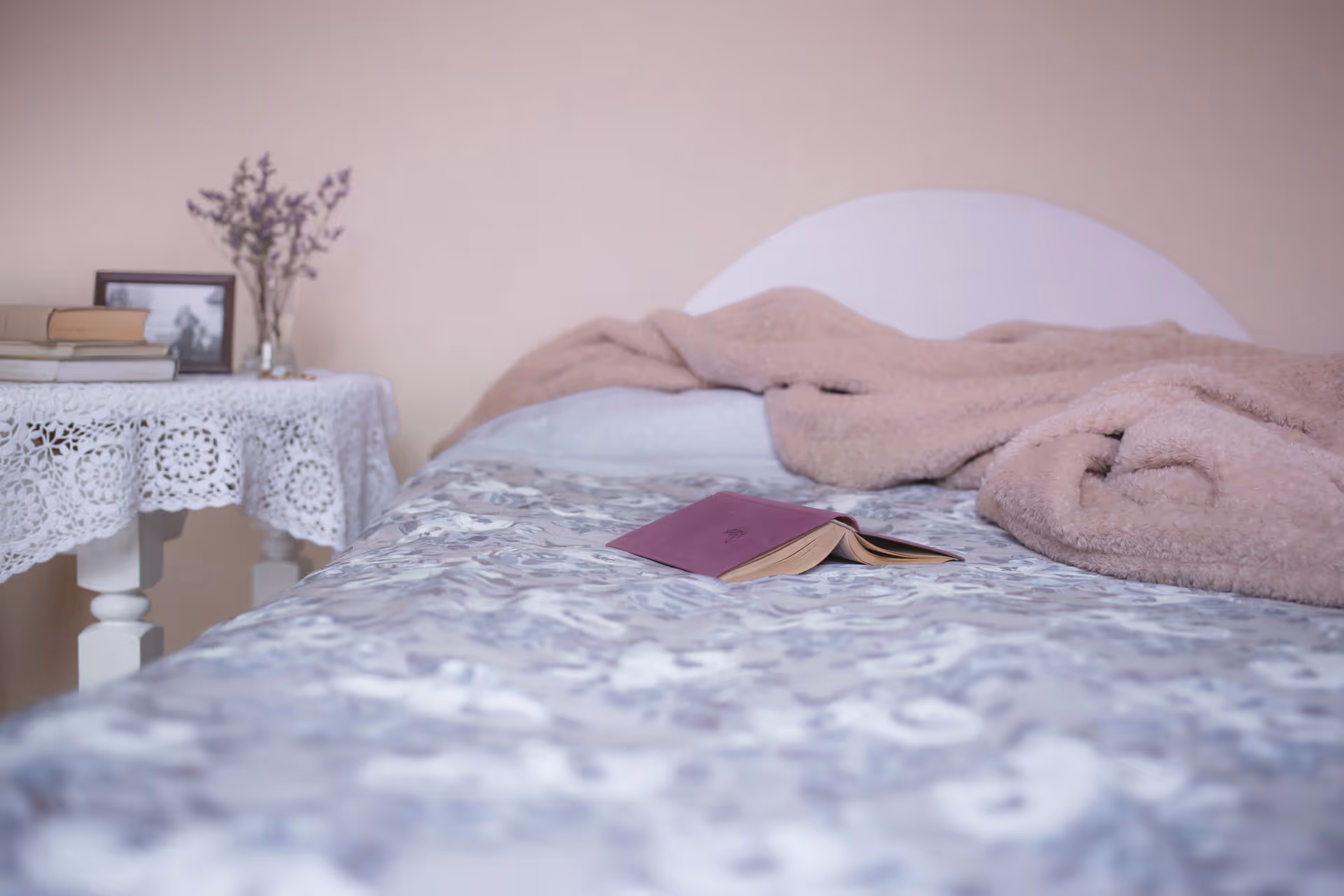 A cozy bedroom with a patterned bedspread, an open book on the bed and a bedside table covered with a lace cloth, books, and a framed photo.