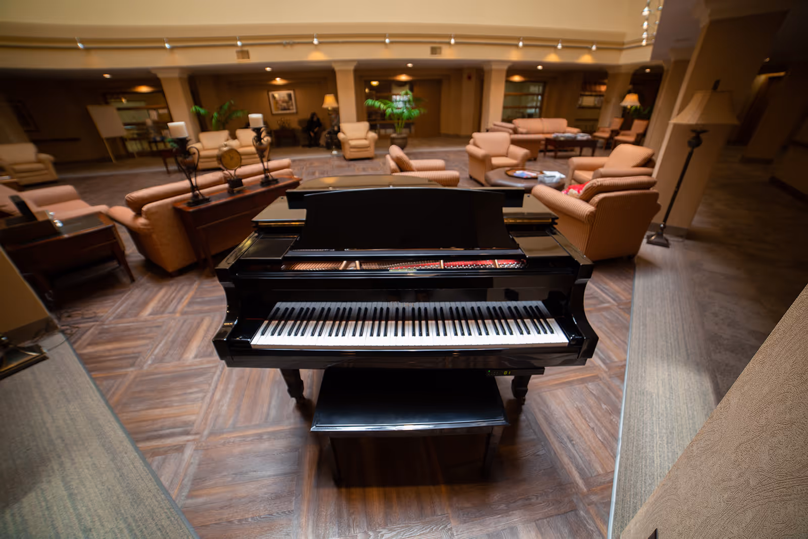 A black grand piano with its lid open is centered in a spacious living room area furnished with multiple beige armchairs and sofas arranged around coffee tables. The room has warm lighting, wooden flooring, and decorative plants, creating a cozy and inviting atmosphere.