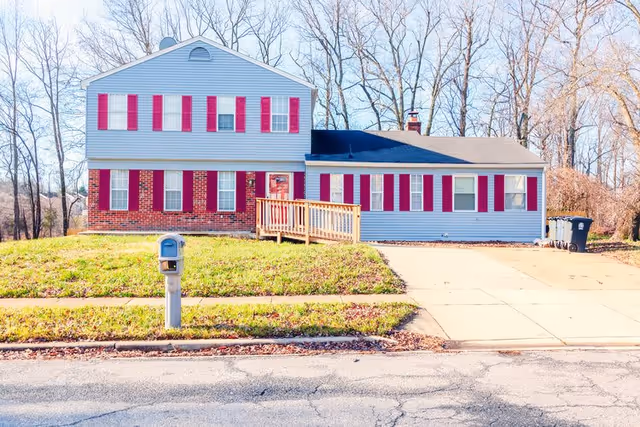 Front exterior view of a two-story residential building with light blue siding and red shutters. There is a wooden ramp leading to the front door, a mailbox on the grassy lawn, and a driveway on the right side. Leafless trees are visible in the background.