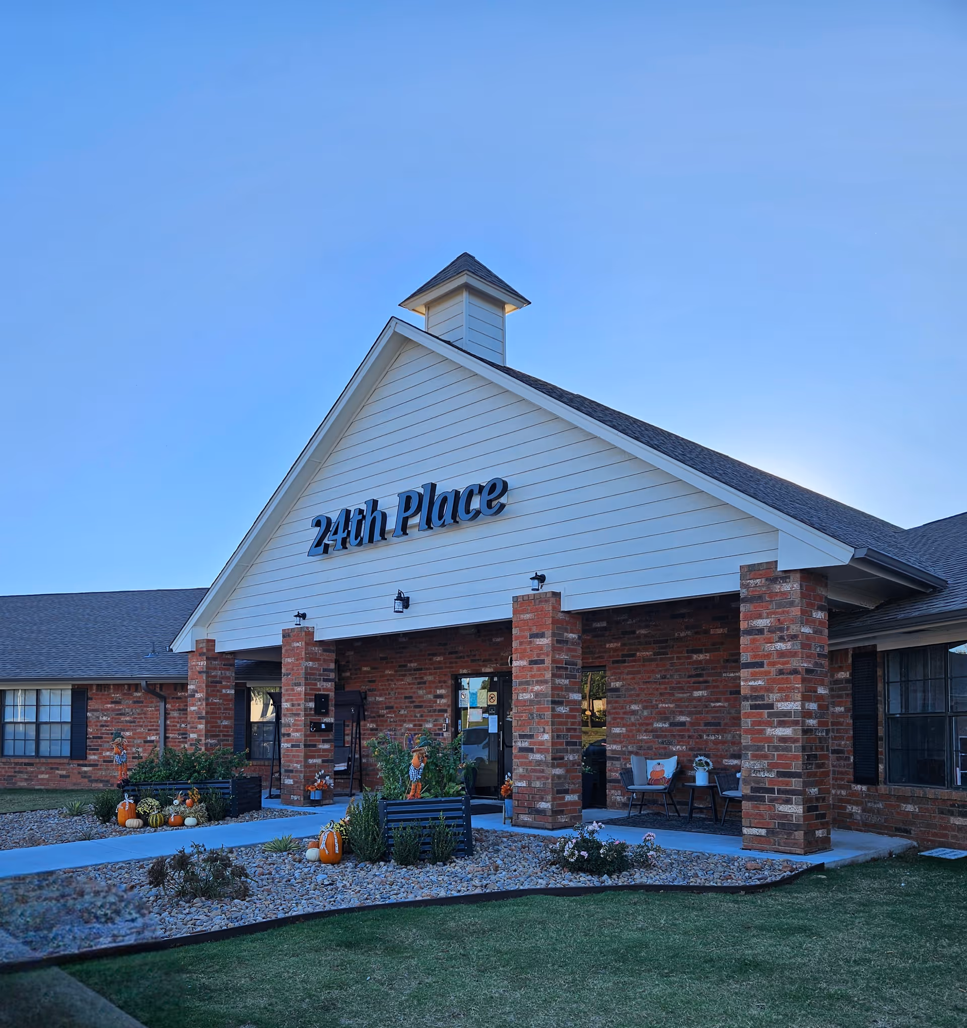 Front entrance of a brick senior living building labeled "24th Place" with a covered porch, landscaping, and pumpkin decorations.