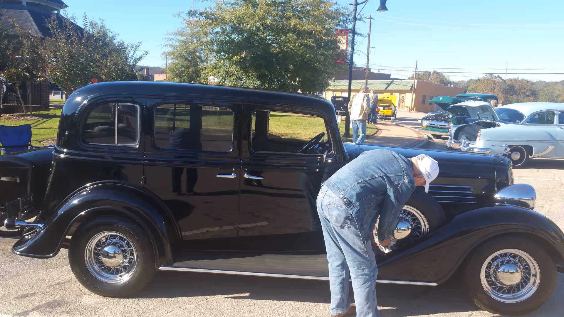 An elderly man wearing a white cap and denim jacket and jeans is polishing the front wheel of a shiny black vintage car parked outdoors. In the background, there are other classic cars with their hoods open and a few people walking around on a sunny day.