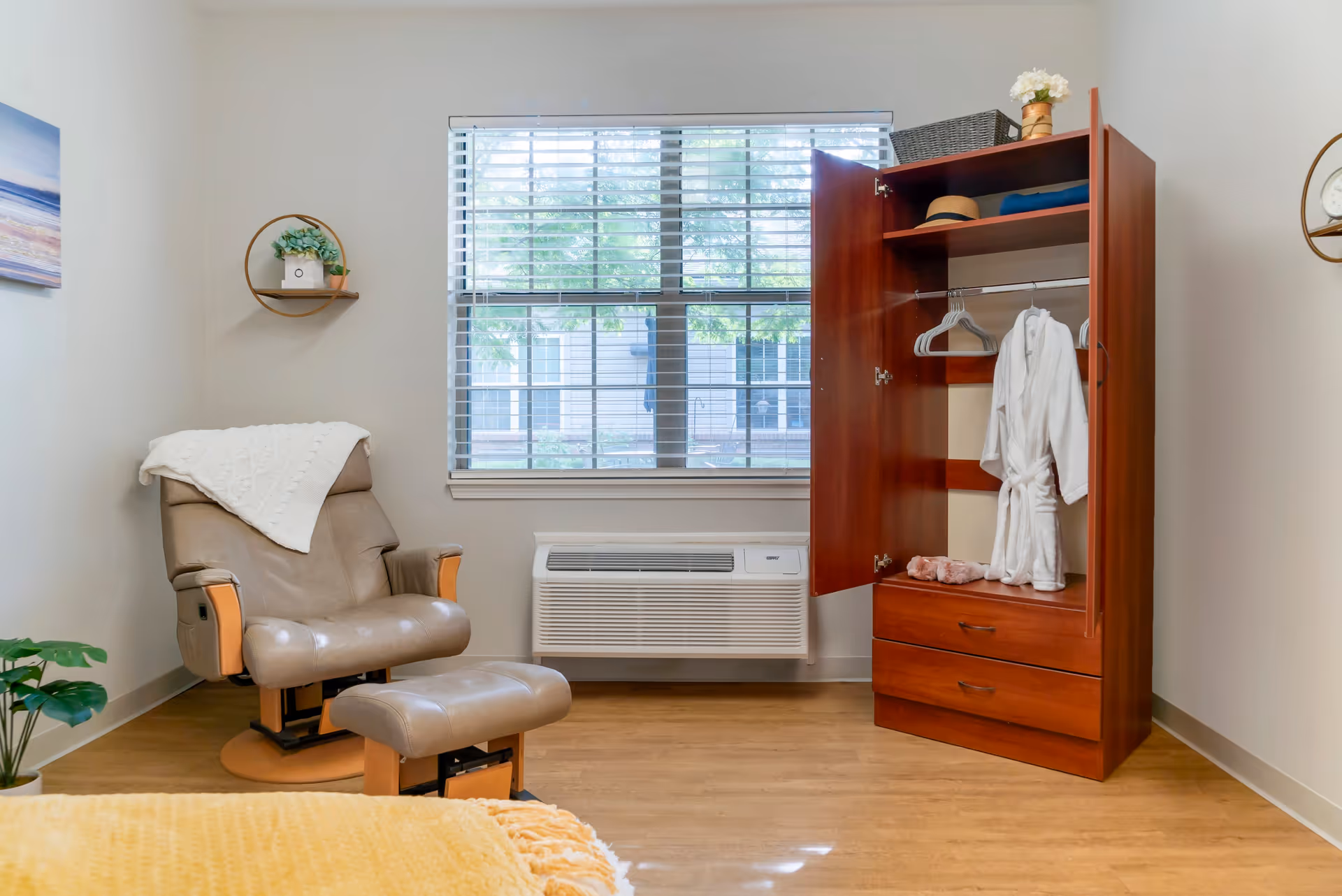 A cozy room with a beige recliner chair and matching footrest, a wooden wardrobe with open doors showing a white robe and some folded clothes, a window with blinds, and light-colored walls and flooring.