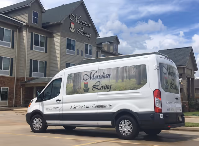 White Meridian Living shuttle van parked in front of the Meridian Living senior care facility building under a partly cloudy sky.