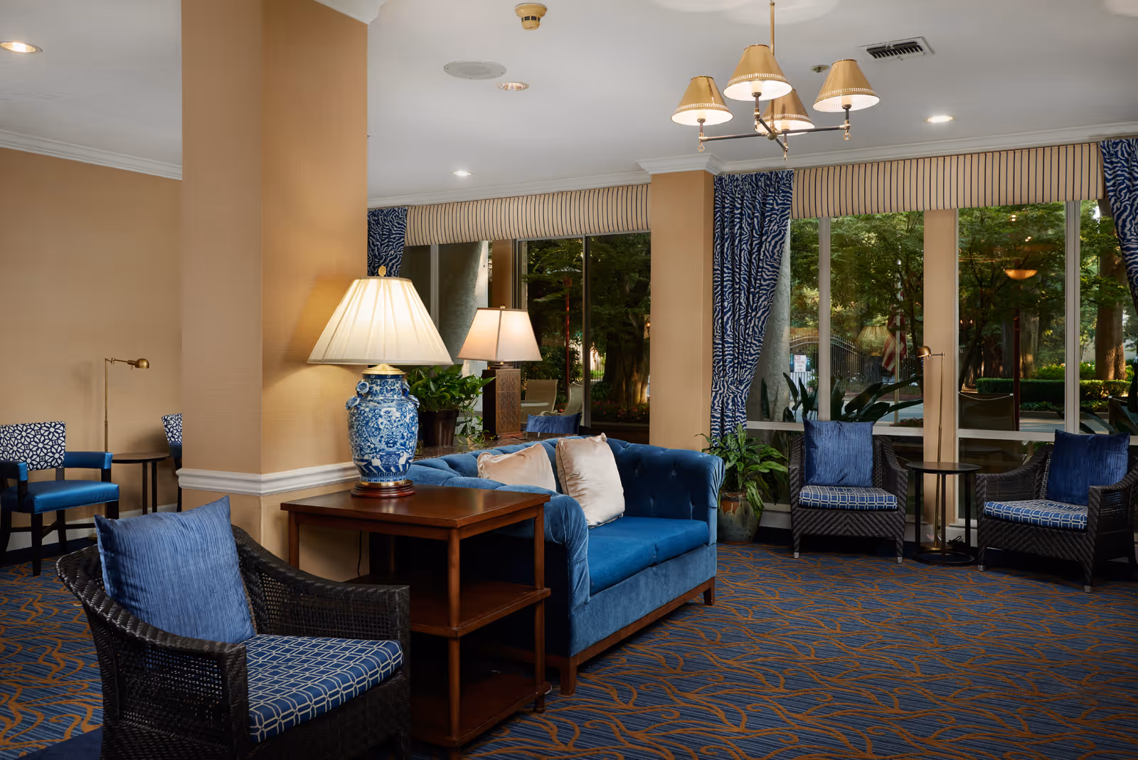 A cozy living room area with blue upholstered chairs and a blue sofa with beige pillows. There are two wooden side tables with lamps, one with a blue and white ceramic base. Large windows with blue patterned curtains let in natural light and show greenery outside. The carpet has a blue and orange pattern, and a chandelier with beige lampshades hangs from the ceiling.