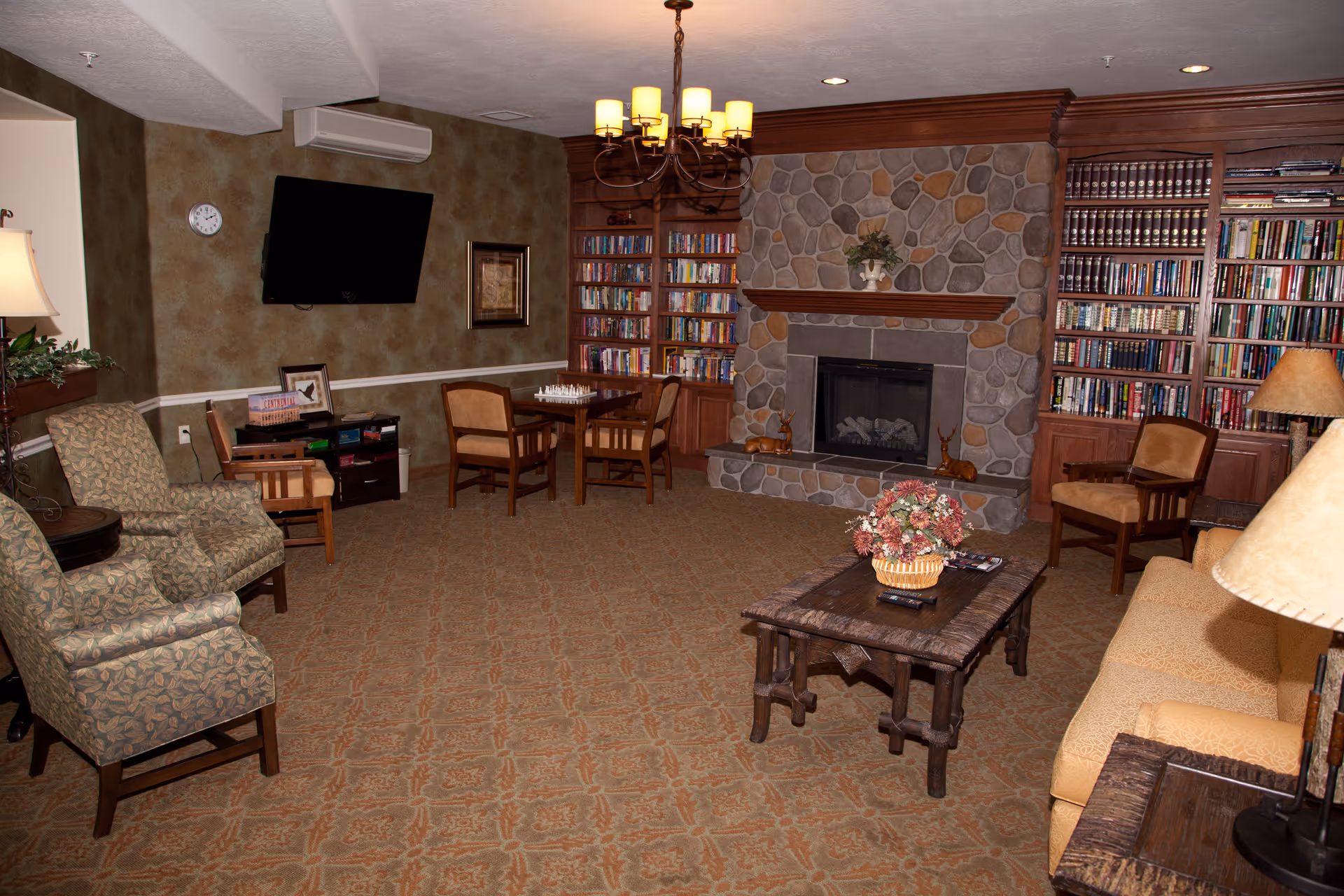A cozy living room area with patterned carpet, a stone fireplace, built-in wooden bookshelves filled with books, a wooden coffee table with a flower arrangement, several upholstered chairs, a sofa, a wall-mounted flat screen TV, a small table with a chessboard, and warm lighting from lamps and a chandelier.