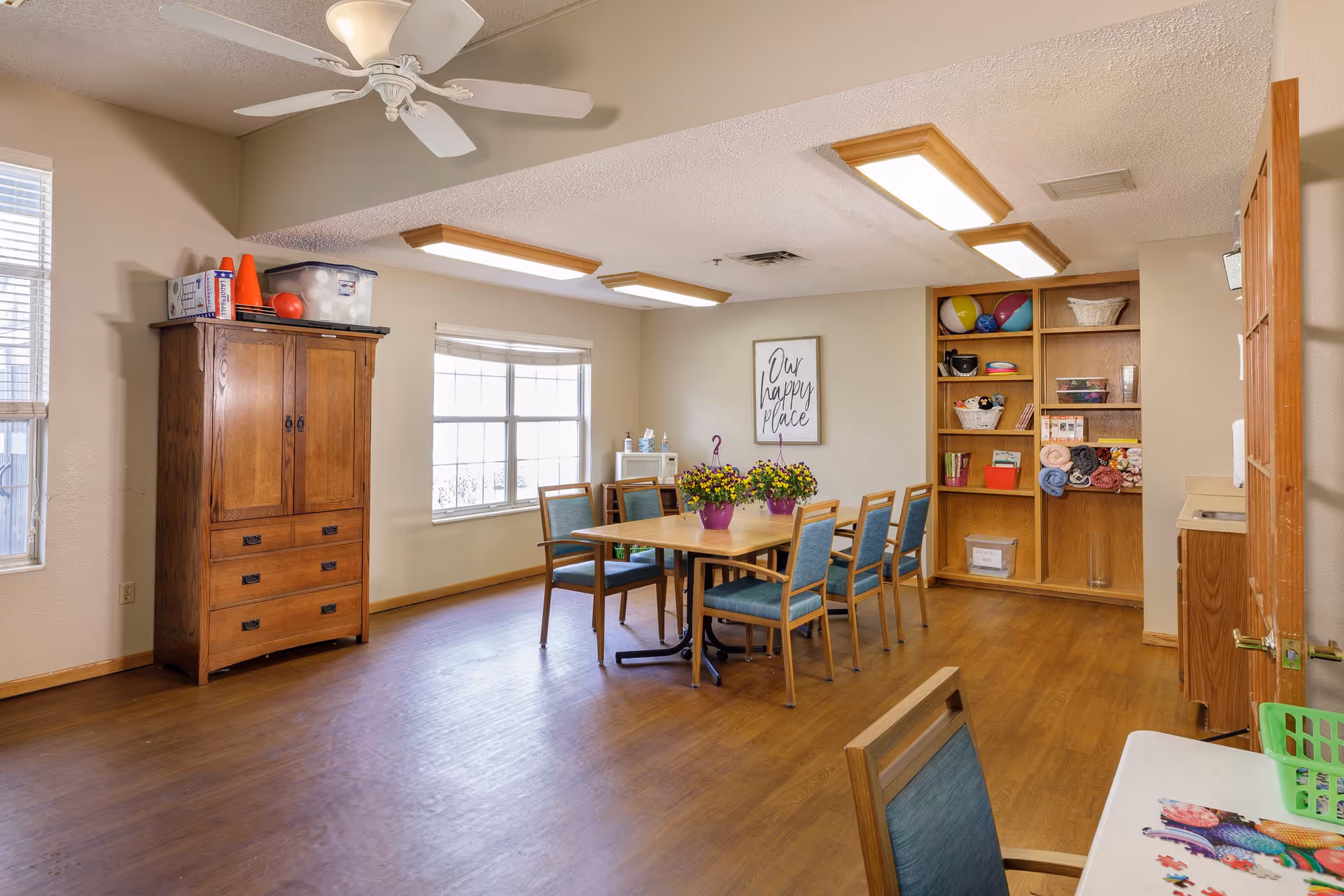 A well-lit room with wooden flooring and beige walls featuring a rectangular table surrounded by six chairs with blue cushions. On the table are two purple pots with yellow flowers. To the left is a wooden cabinet with various items on top, including orange cones and a plastic container. A large window with blinds lets in natural light. On the far wall is a framed sign that reads 'Our happy place'. To the right, there is a wooden shelving unit holding colorful balls, baskets, books, and folded blankets. A ceiling fan and fluorescent lights are mounted on the ceiling.