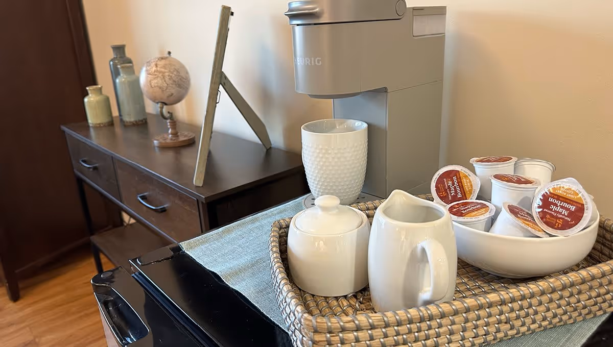 A coffee station setup with a Keurig coffee maker, a white textured mug, a white ceramic sugar bowl, a white ceramic creamer pitcher, and a bowl filled with maple bourbon flavored coffee pods. In the background, there is a dark wooden sideboard with decorative items including two vases and a small globe.