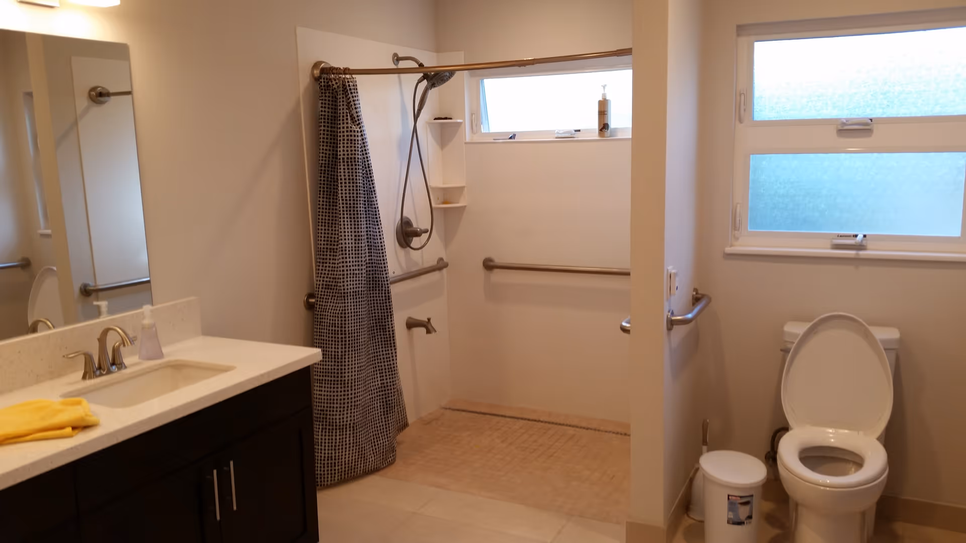 A clean and accessible bathroom featuring a sink with a countertop and a mirror on the left, a walk-in shower with a handheld showerhead and a black and white checkered shower curtain in the center, and a toilet with the lid open on the right. The bathroom has grab bars installed near the shower and toilet, and two frosted windows providing natural light.