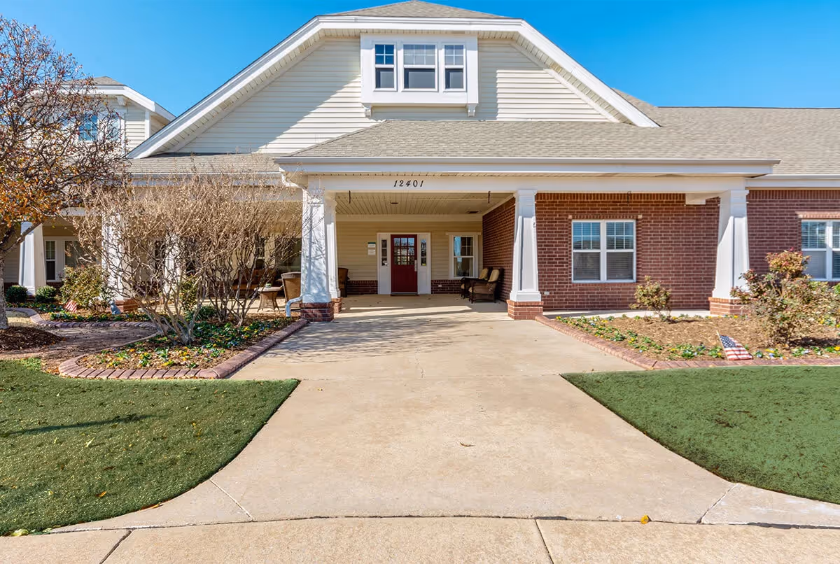 Front exterior view of Dorset Place Assisted Living building with a covered entrance, brick and siding facade, windows, and landscaped garden beds on either side of the driveway.