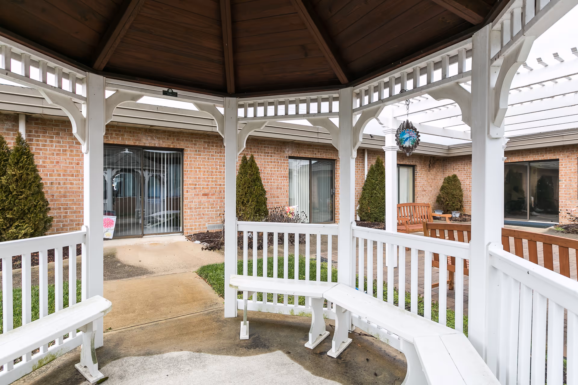 View from inside a white wooden gazebo with built-in benches, looking out onto a courtyard with brick walls, sliding glass doors, small evergreen shrubs, and wooden benches under a white pergola.