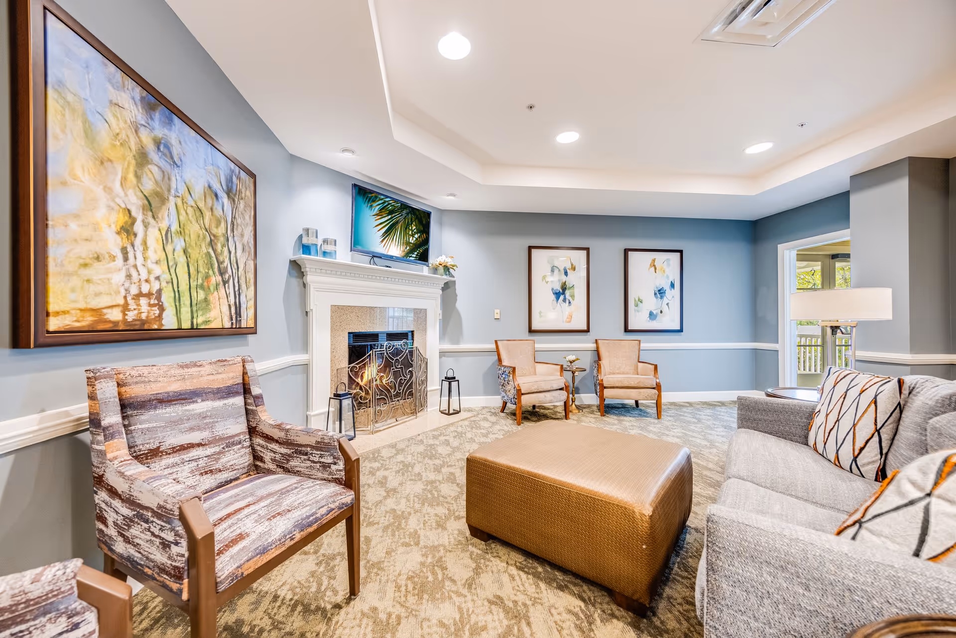 A cozy living room with light blue walls, a fireplace with a decorative screen, a TV mounted above the fireplace, and various seating options including a patterned armchair, two wooden chairs with cushions, a large ottoman, and a gray sofa with patterned pillows. The room is decorated with abstract paintings and has a carpeted floor.