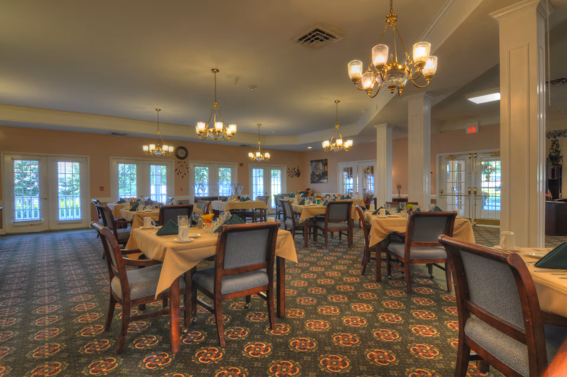A spacious dining room with multiple tables covered in beige tablecloths, each set with glasses, napkins, and plates. The room features patterned carpet, several chandeliers hanging from the ceiling, large windows and glass doors letting in natural light, and white columns along one side.