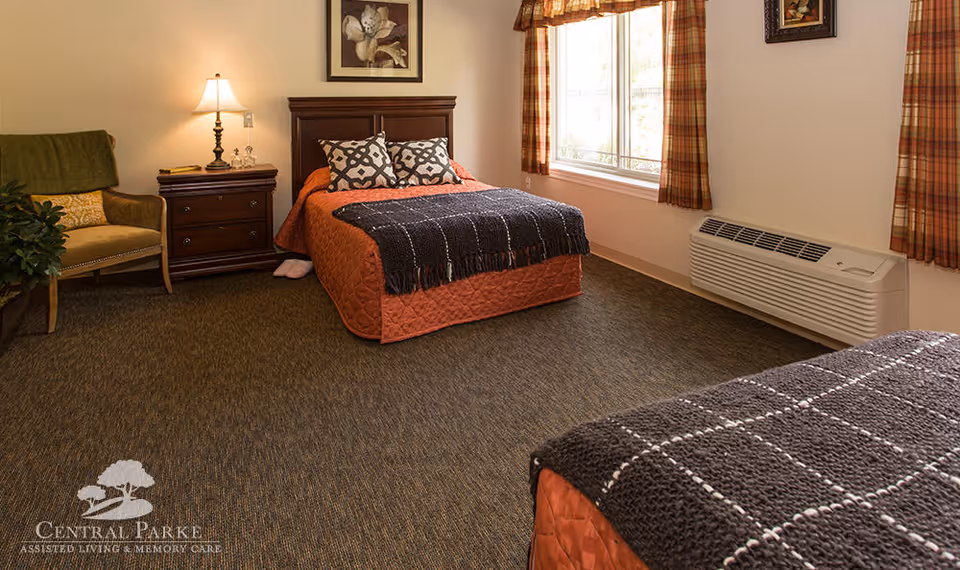A cozy bedroom in an assisted living facility featuring two beds with orange bedspreads and dark gray knitted throws. There is a wooden nightstand with a lamp and decorative items between the beds. A green armchair with a yellow pillow is placed in the corner next to a plant. The room has carpeted flooring, a large window with plaid curtains, and a wall-mounted air conditioning unit below the window. A framed flower picture hangs above the nightstand.
