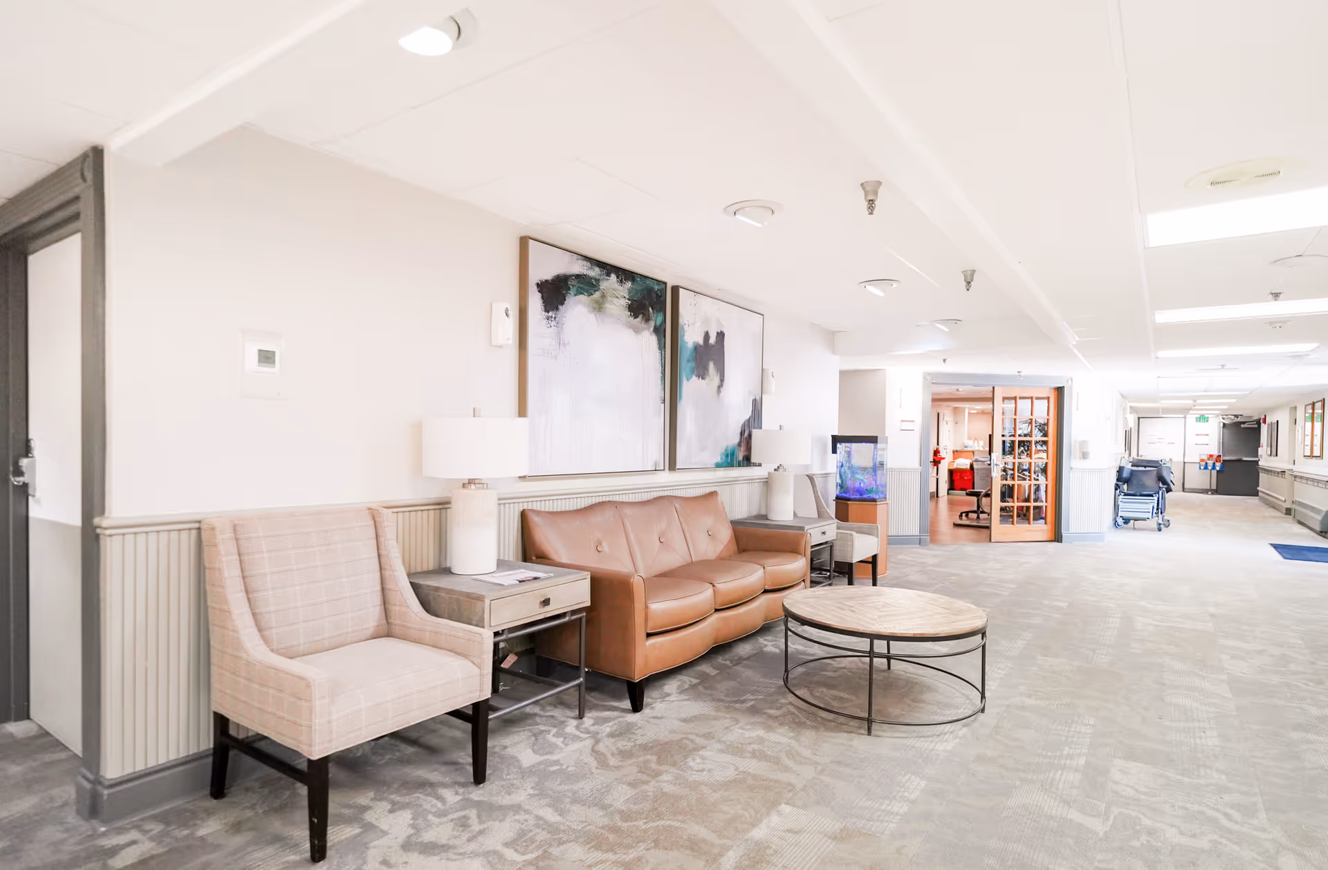 A bright and spacious hallway seating area in a senior living facility featuring a tan leather sofa, a beige upholstered armchair, two side tables with lamps, a round coffee table, abstract wall art, and a fish tank. The hallway extends into other rooms and has a wheelchair visible in the distance.