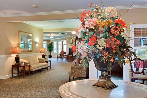 A spacious and warmly lit common area in an assisted living facility featuring a large floral arrangement in a decorative vase on a round table in the foreground. In the background, there is a beige sofa with a side table and lamp, framed artwork on the wall, and additional seating with chairs and tables further down the carpeted hallway.