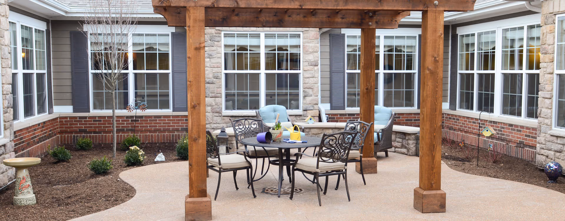 Outdoor patio area at Bickford of Gurnee featuring a wooden pergola with a round metal table and four cushioned chairs underneath. The patio is surrounded by a garden bed with small shrubs and decorative items, and the building's windows and brick exterior are visible in the background.