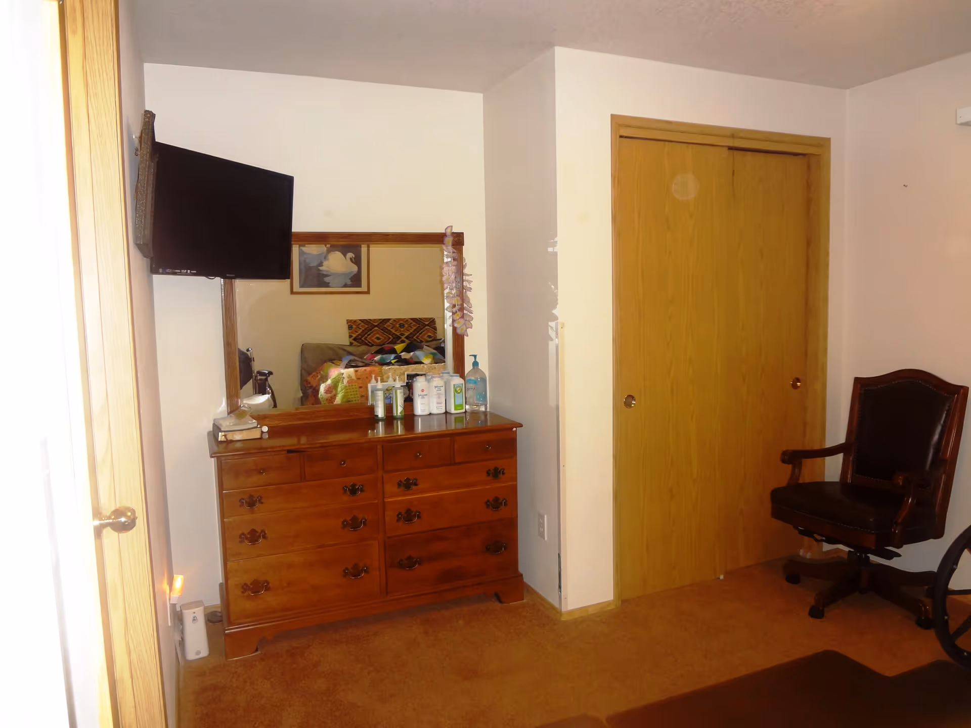 A bedroom corner featuring a wooden dresser with a mirror above it, various bottles and a hand sanitizer on top, a wall-mounted flat-screen TV to the left, a wooden sliding closet door, and a dark leather office chair on wheels. The room has beige walls and carpeted floor.