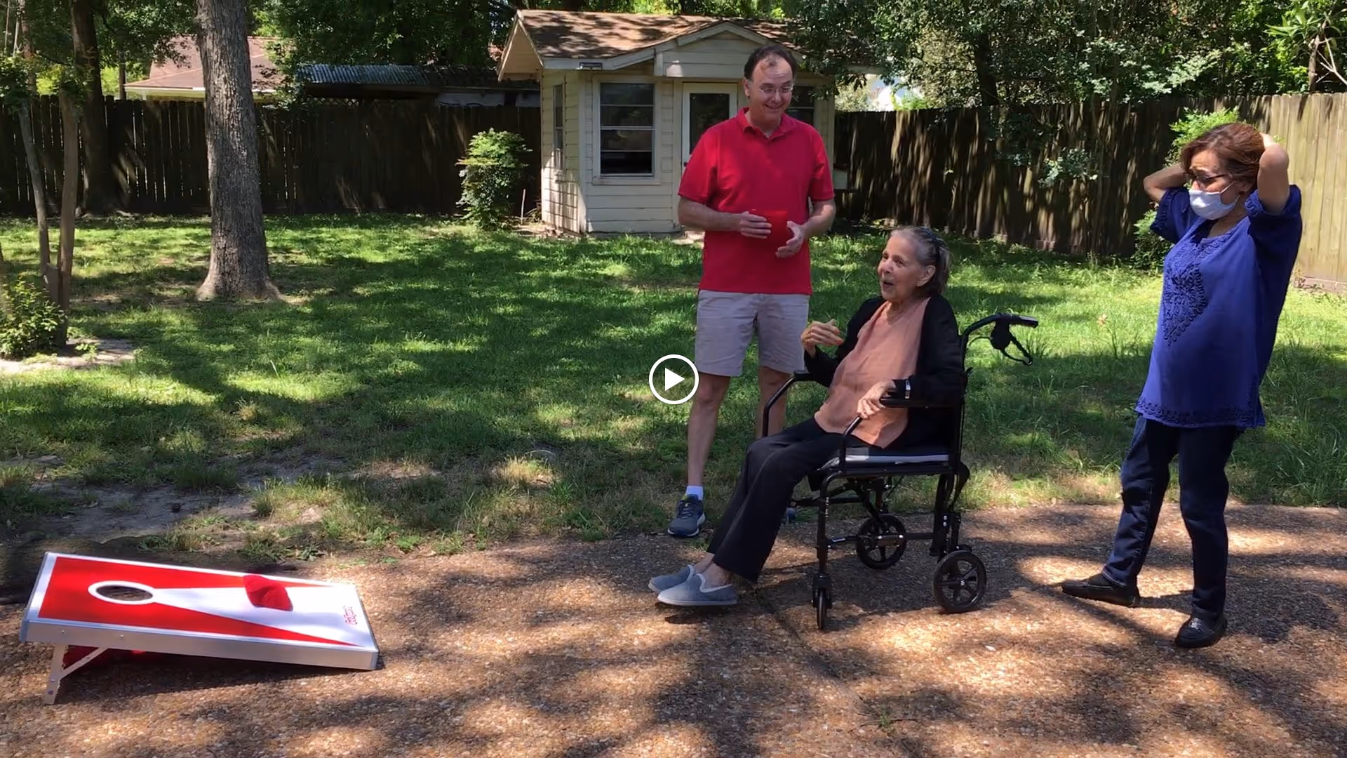 Three people in a backyard setting with green grass and trees. One elderly woman is seated in a wheelchair, engaging with a man in a red shirt and beige shorts standing nearby. Another woman wearing a blue top and a face mask stands with her hands behind her head. A cornhole game board is set up on the ground in front of them. A small shed and wooden fence are visible in the background.