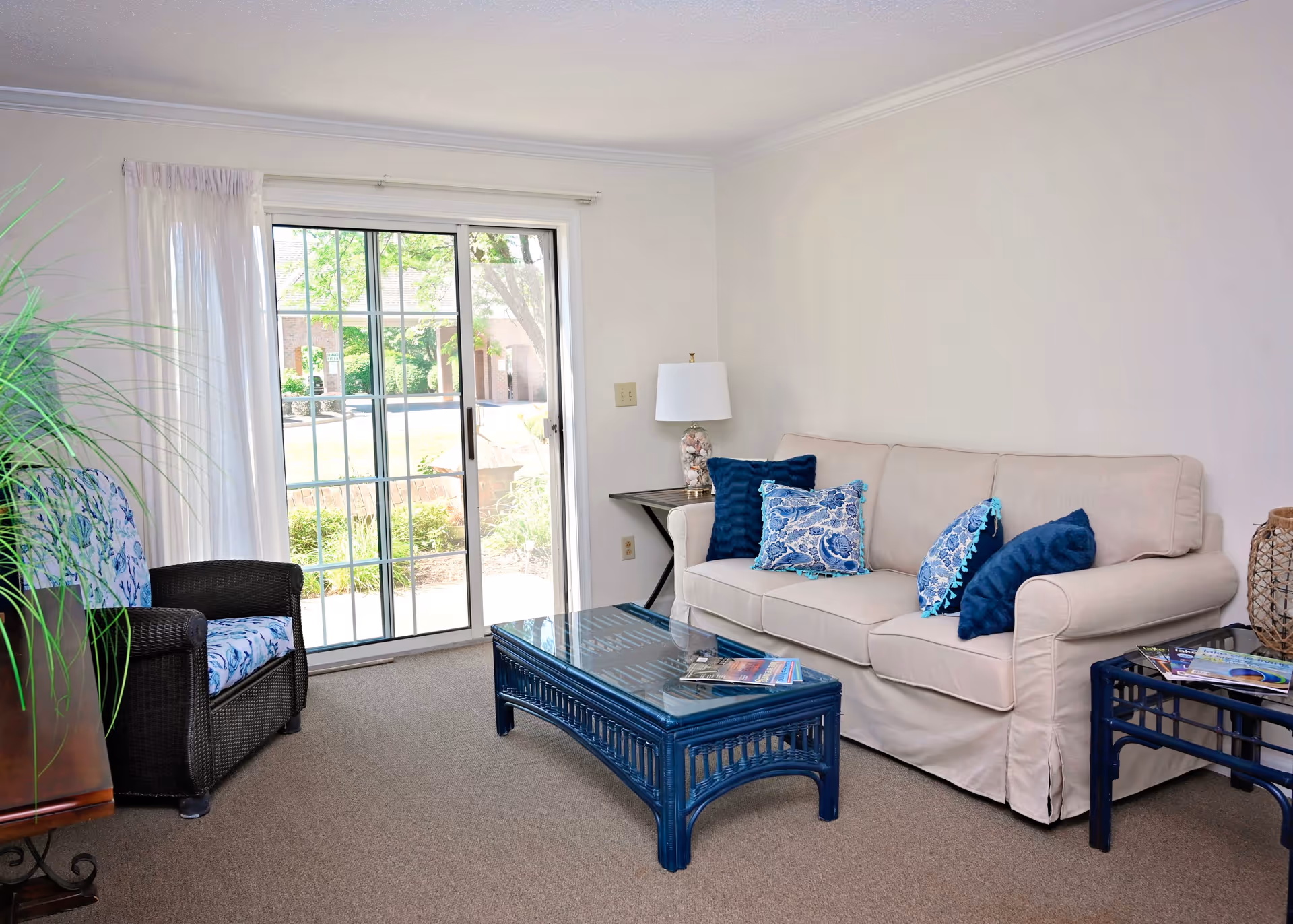 A bright living room with a beige sofa adorned with blue and patterned throw pillows, a glass-top blue wicker coffee table, a matching side table with magazines, a black wicker armchair with a blue patterned cushion, a table lamp on a small side table, and a sliding glass door with sheer white curtains leading to an outdoor area.