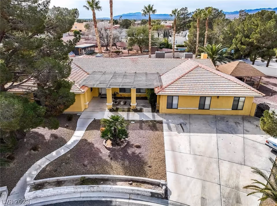 Aerial view of a single-story yellow building with a tiled roof, surrounded by trees and desert landscaping. There is a curved concrete walkway leading to the entrance, a small circular garden with a palm tree in front, and a large concrete driveway on the right side. The background shows more trees, houses, and distant mountains under a clear sky.
