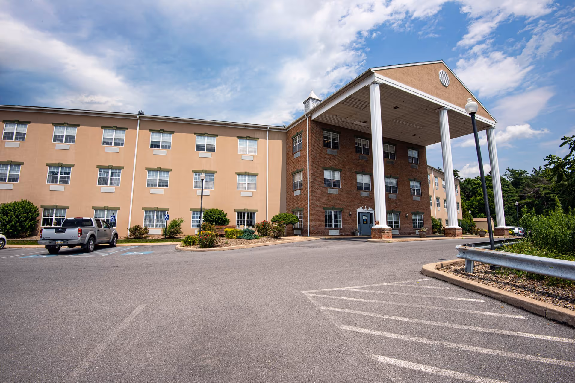 Exterior view of a three-story nursing and rehabilitation center building with a large covered entrance supported by four white columns. The building has beige and brick walls with multiple windows. There is a parking lot in front with a few cars parked and some greenery around the building under a partly cloudy sky.
