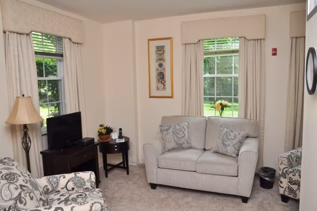 A cozy living room with a beige loveseat, patterned armchairs, a small round side table with a plant and phone, a floor lamp, and a flat-screen TV on a wooden stand. Two windows with beige curtains let in natural light and show greenery outside. A framed artwork hangs on the wall between the windows.