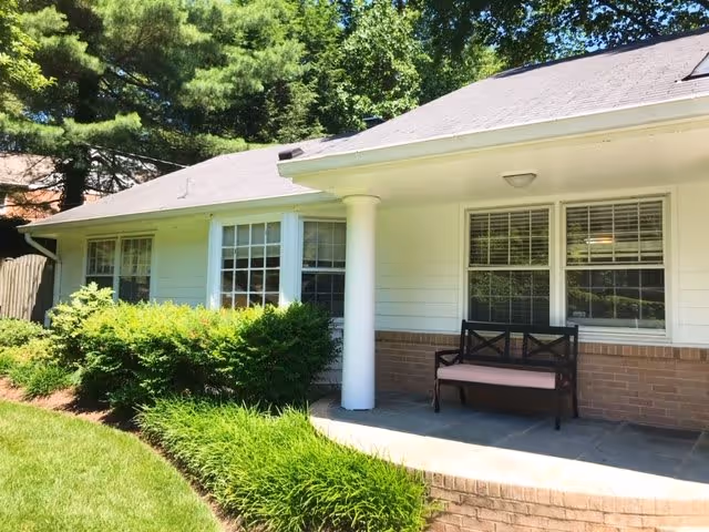 Exterior view of a single-story building with white siding and brick accents, featuring a small covered porch with a black bench. The porch is supported by a white column and surrounded by green bushes and grass, with trees in the background.