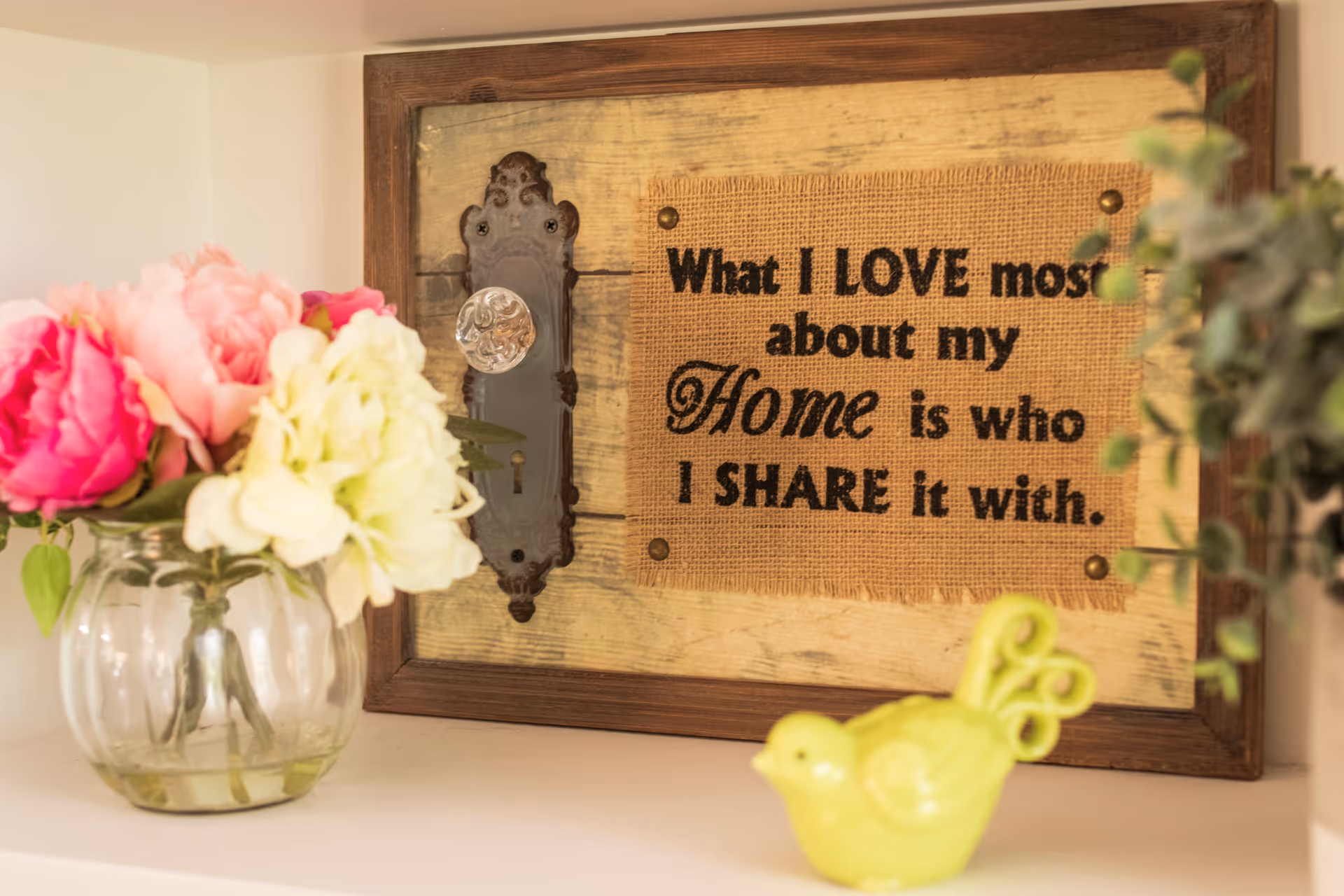 A decorative wooden plaque with a vintage door handle and a burlap patch that reads 'What I LOVE most about my Home is who I SHARE it with.' Next to the plaque is a glass vase with pink and white flowers and a small green ceramic bird figurine.