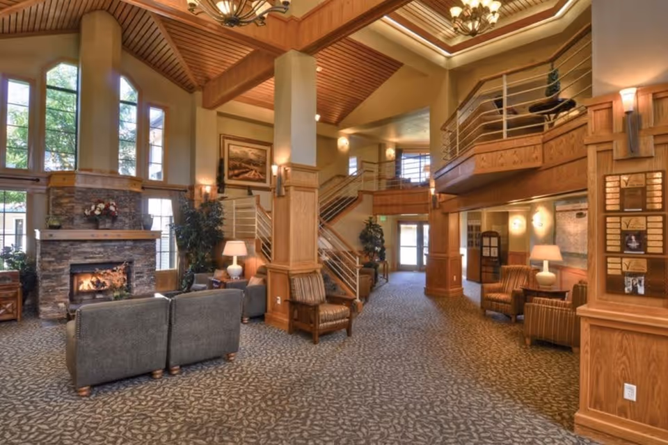 A spacious and warmly lit senior living facility common area with high wooden ceilings, large windows, a stone fireplace with a fire burning, comfortable seating including sofas and armchairs, carpeted floor, and a staircase leading to an upper level with railings.