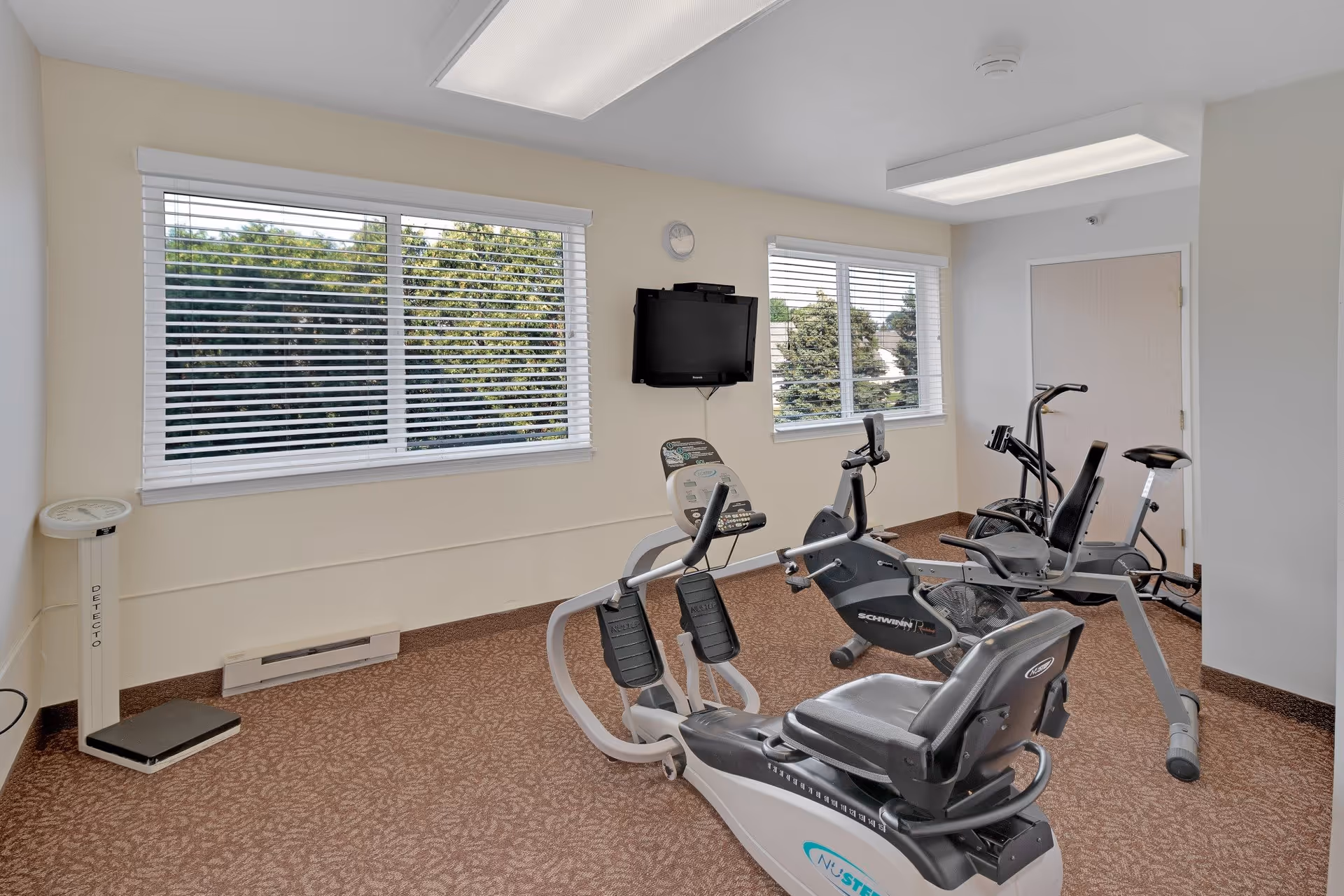 A small exercise room with three stationary exercise bikes, a wall-mounted TV, two windows with white blinds showing trees outside, a wall clock, and a standing scale in the corner. The room has beige walls and carpeted floor.