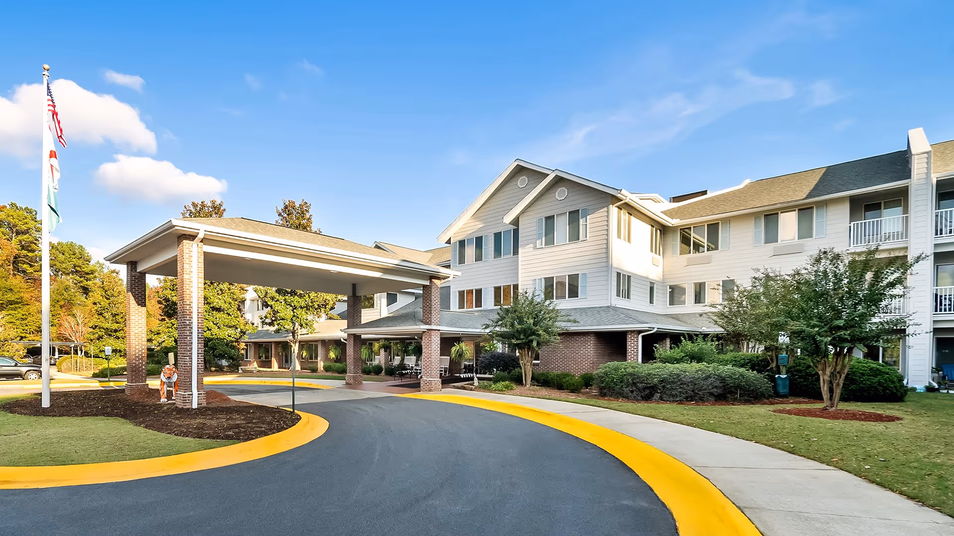 Exterior view of Monarch Estates, a multi-story senior living facility with a covered entrance driveway, surrounded by landscaped greenery and trees under a clear blue sky.