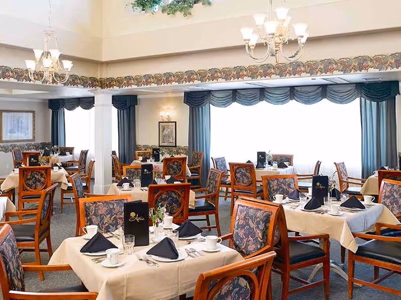A well-lit dining room with multiple tables covered in beige tablecloths, each set with white cups, glasses, silverware, and black folded napkins. The chairs have wooden frames with floral-patterned upholstery. Large windows with blue curtains allow natural light to fill the room. Decorative chandeliers hang from the ceiling, and framed artwork adorns the walls.